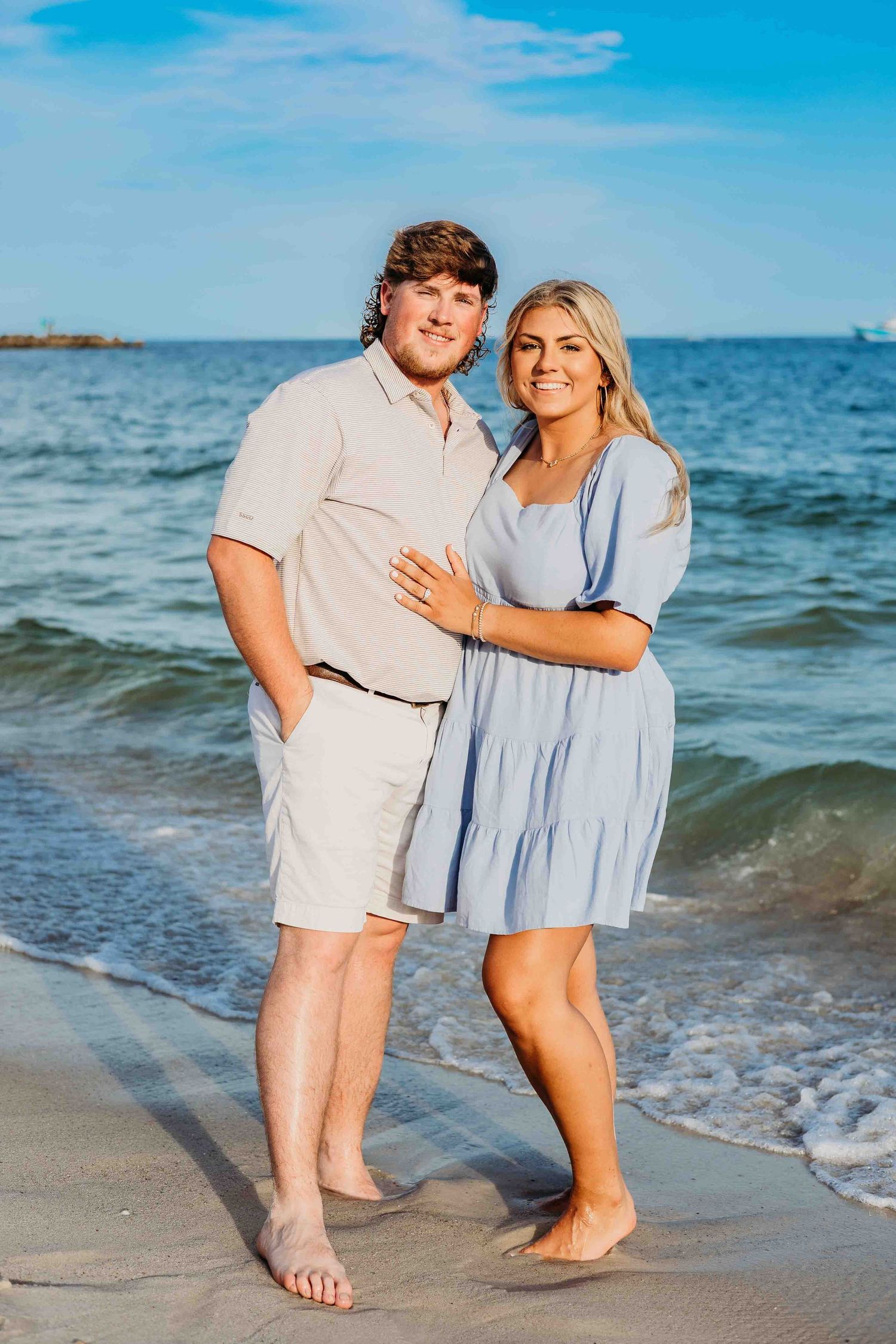 Romantic couple walking along the beach shoreline in coordinated white and blue outfits at sunset.