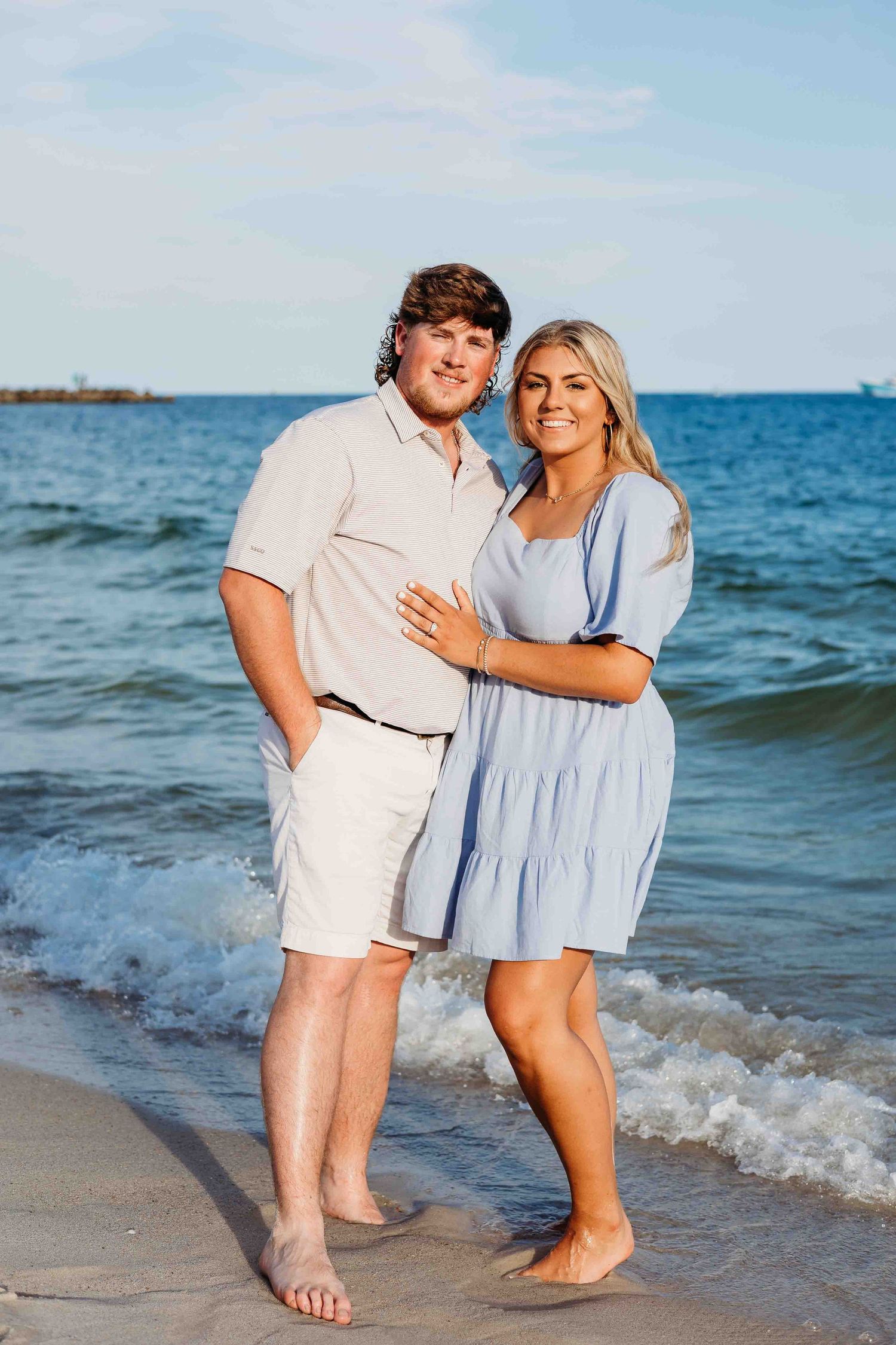 Happy couple embracing on a sandy beach with gentle waves and blue ocean in the background.