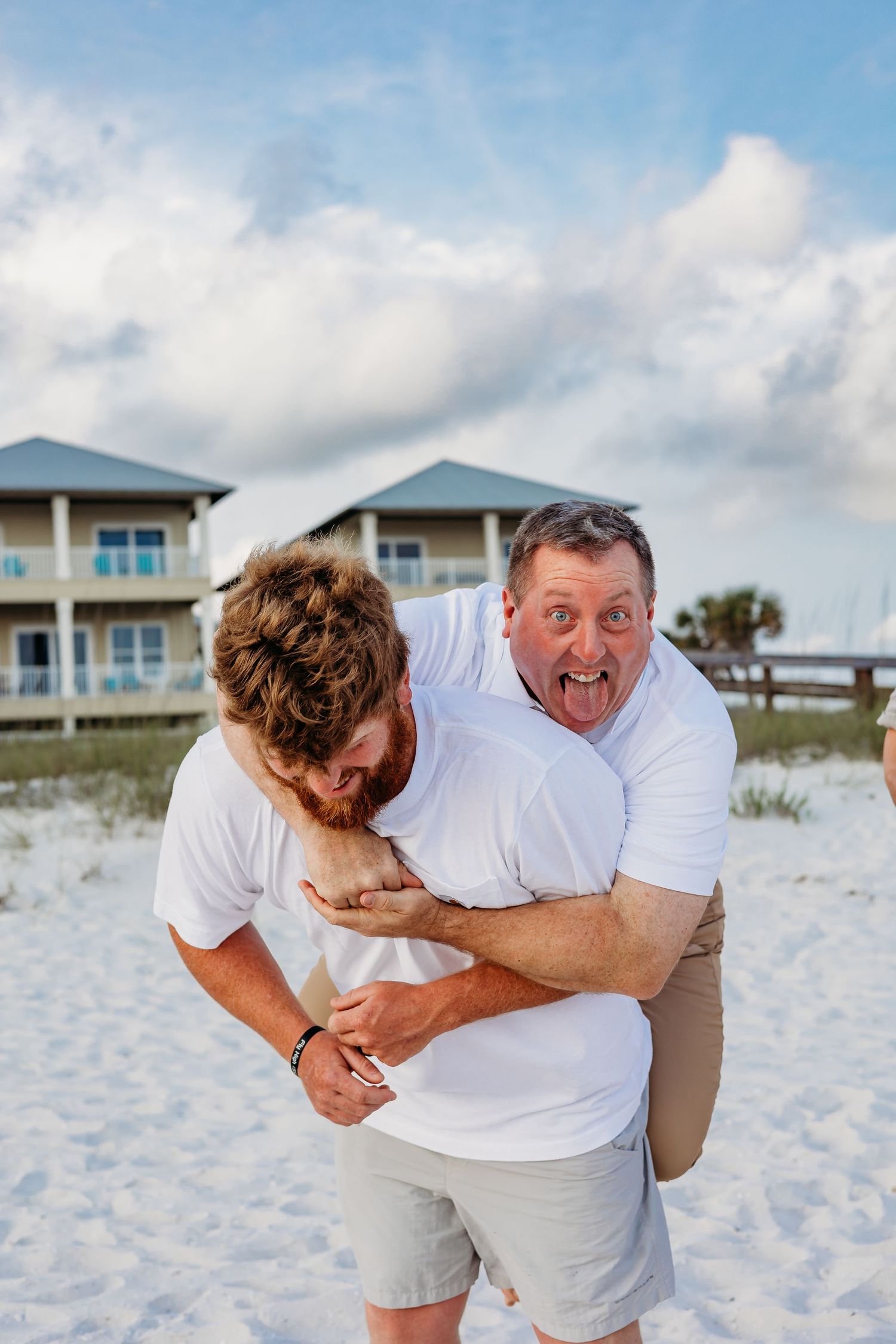 Two people playfully horsing around on a white sandy beach with beach houses visible behind them.