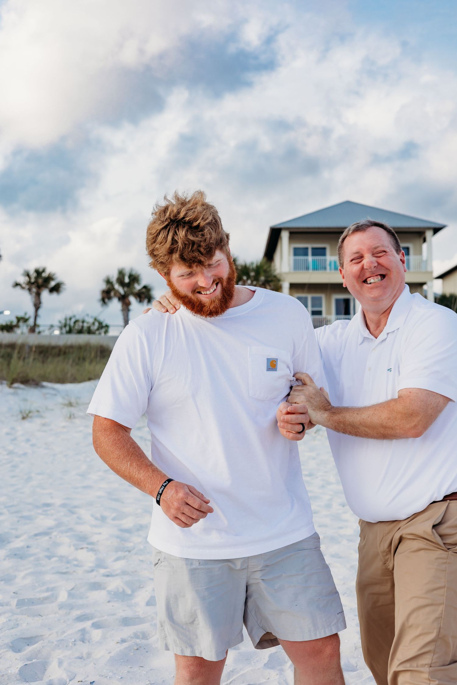 Two people in white shirts share a lighthearted moment on the beach with coastal homes in the background.