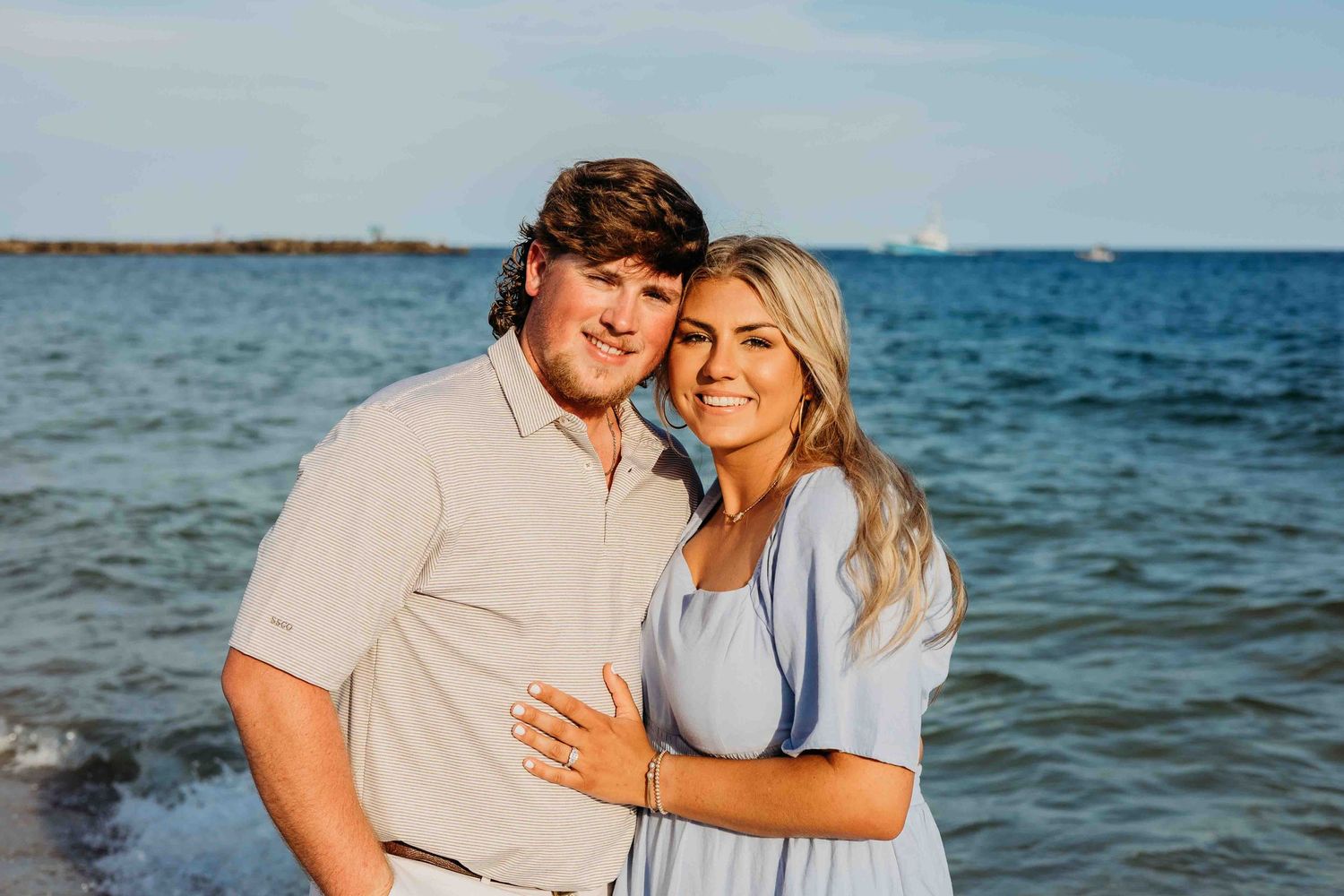Couple in light summer clothing posing for romantic beach photos with ocean waves behind them.