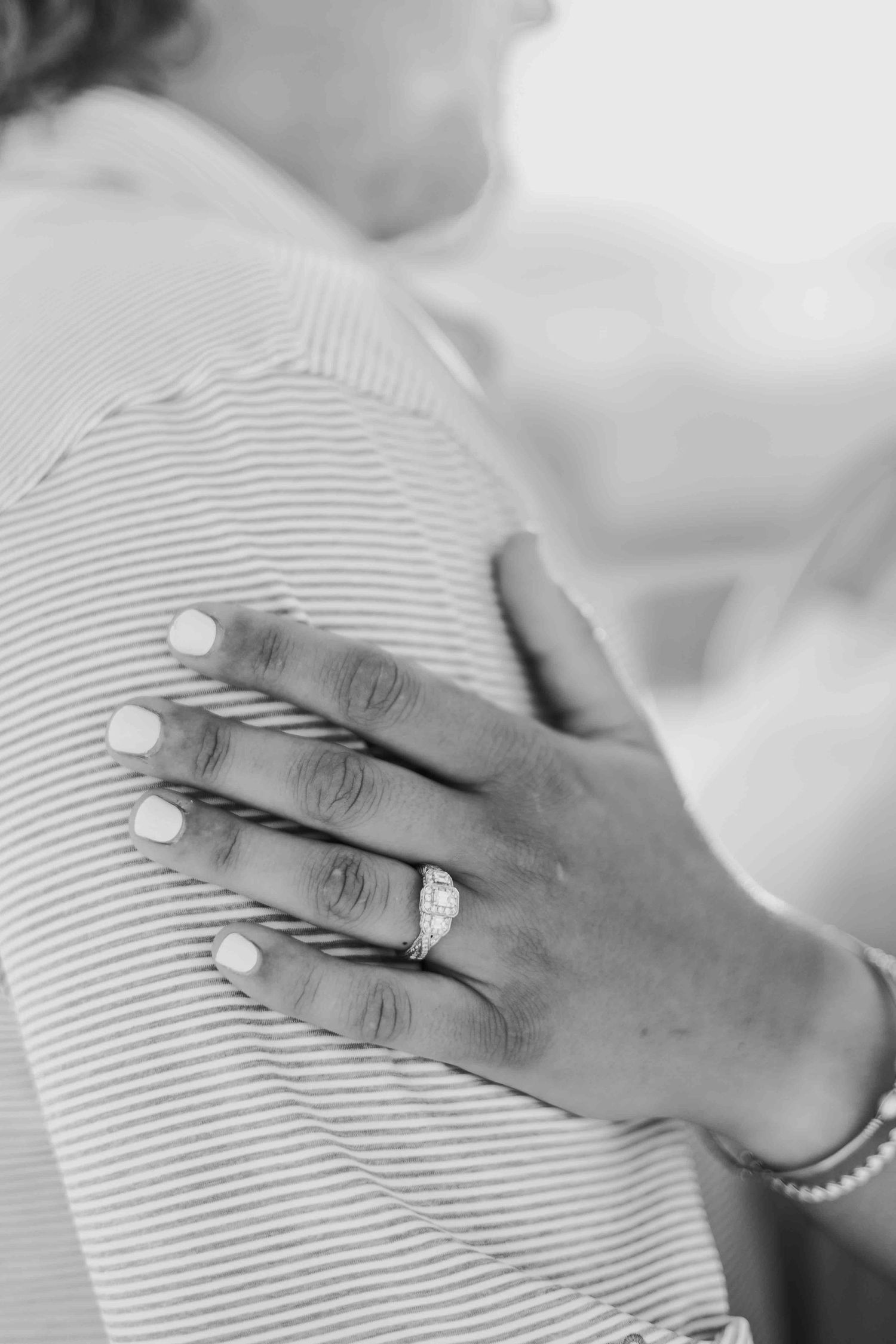 Black and white detail shot of a hand with engagement ring and bracelet against textured fabric.
