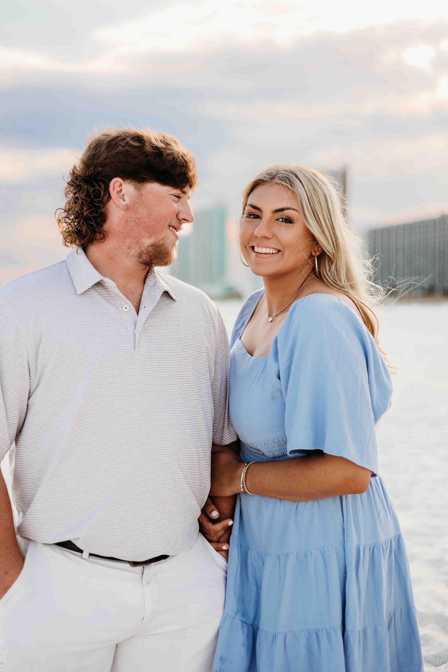 Couple in white and blue outfits share romantic moments on a beach with city buildings in background.