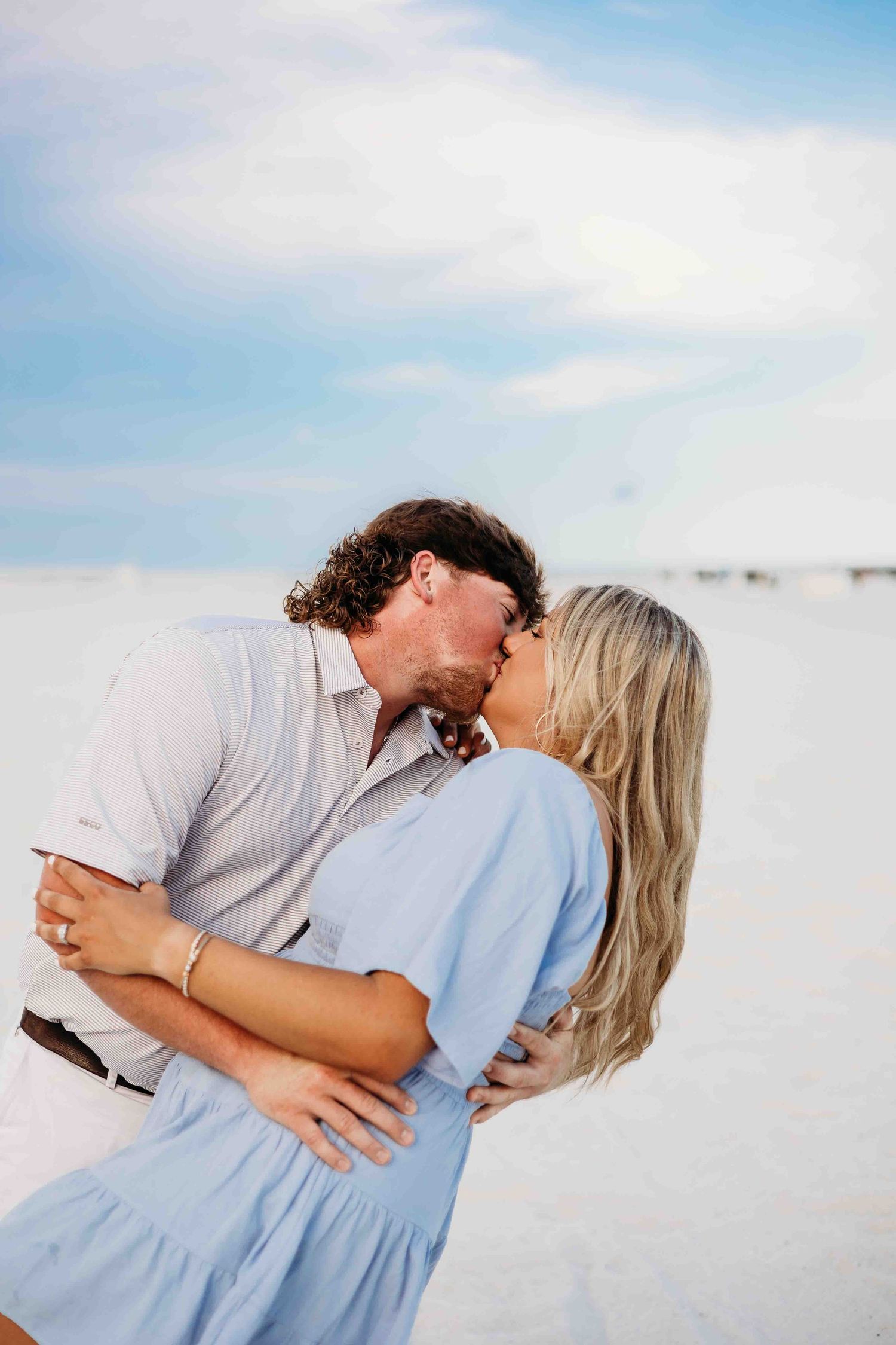 Romantic couple embracing on a white sandy beach with soft blue sky in the background.