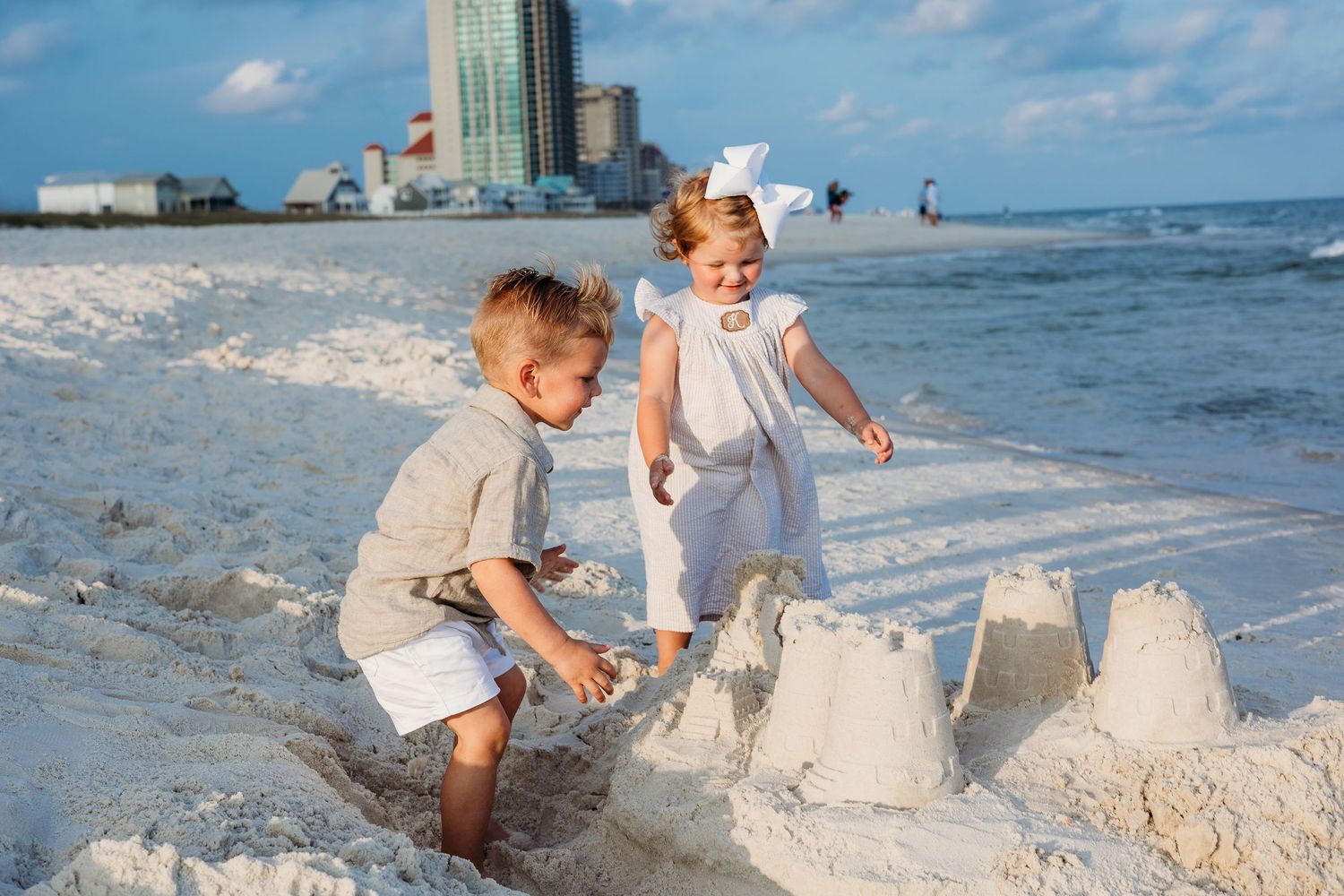 Children building sandcastles on a sunny beach with high-rise buildings in the background.