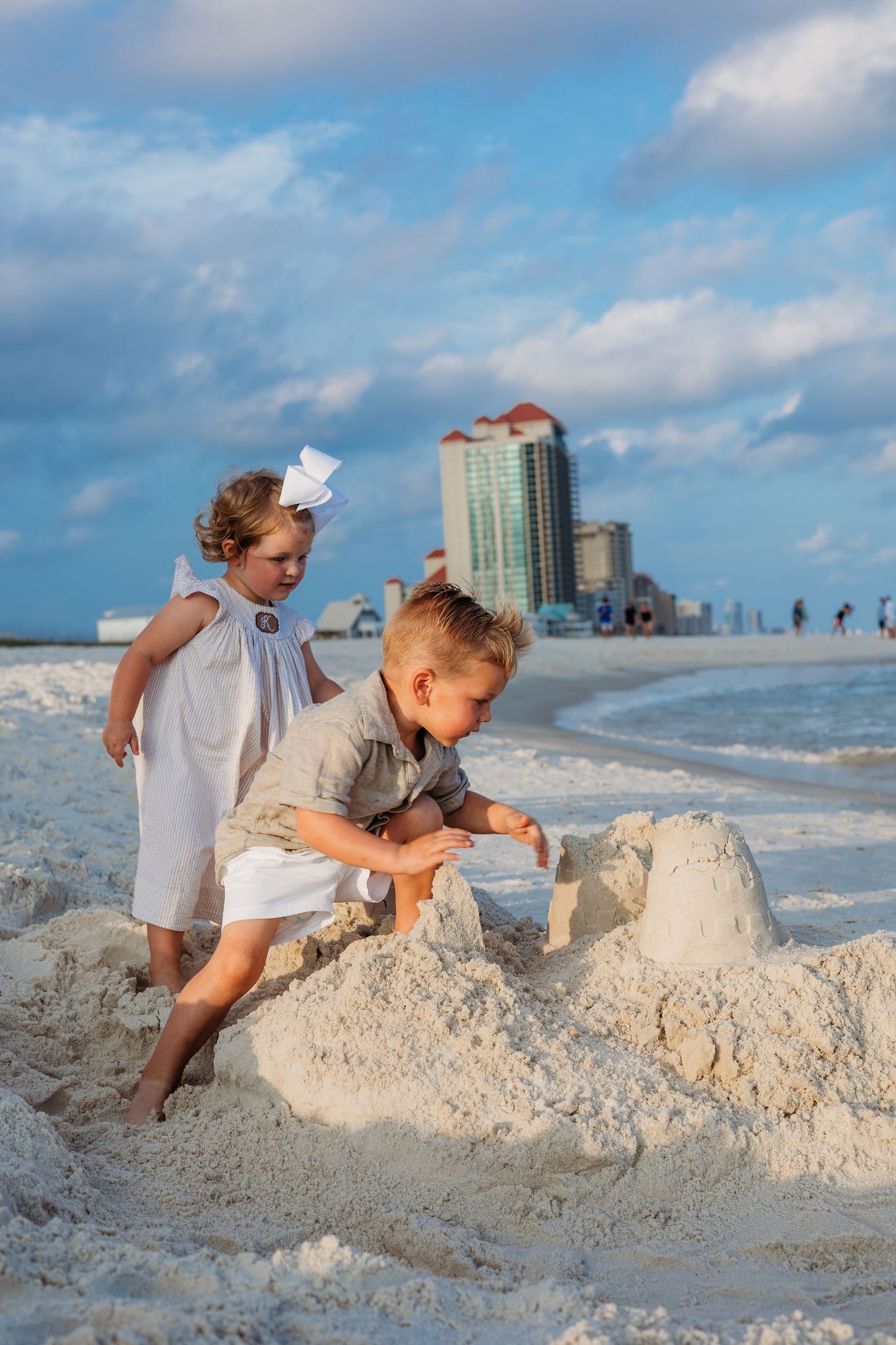 Kids playing in white sand on a beach with coastal buildings visible in the distance.