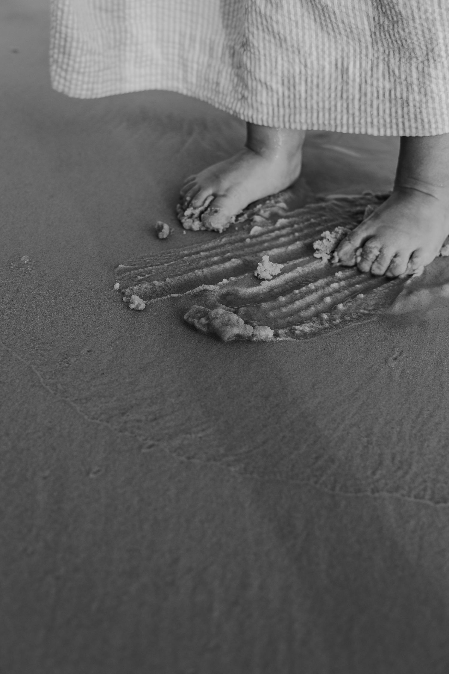 Black and white close-up of small feet next to seashells arranged on dark sand.