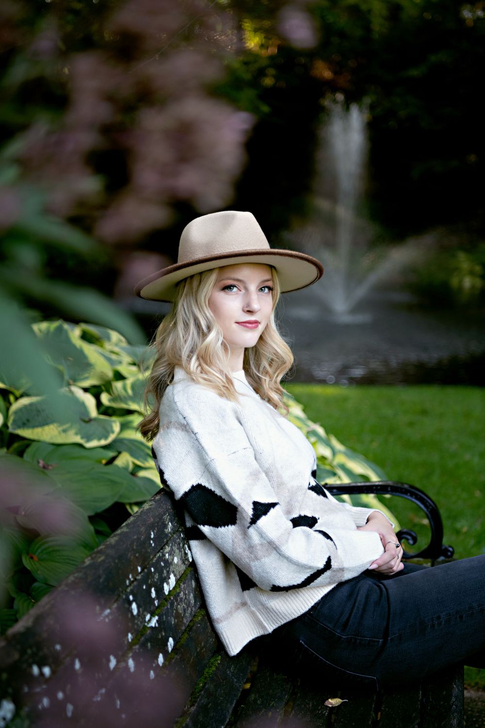 Stylish portrait of someone in a beige hat and white sweater sitting on a bench in a garden setting.