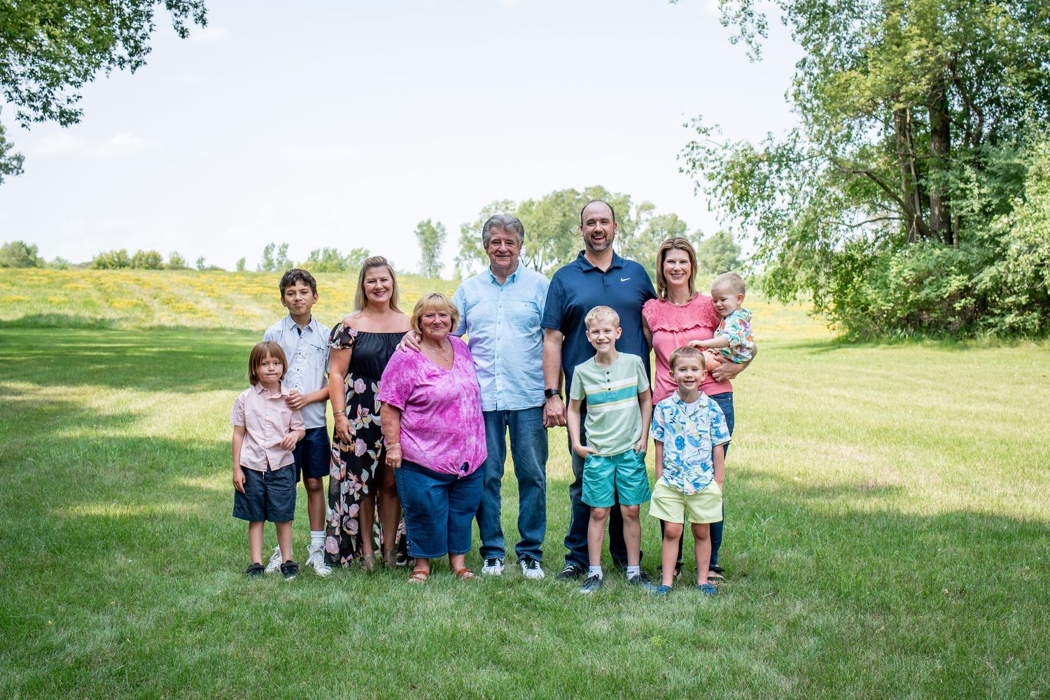 Family group portrait taken outdoors in sunny green field with trees in background.