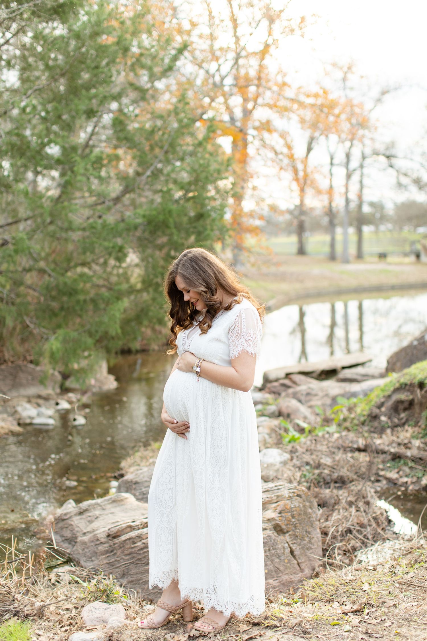 A woman in flowing white dress cradles her baby bump by a scenic stream.