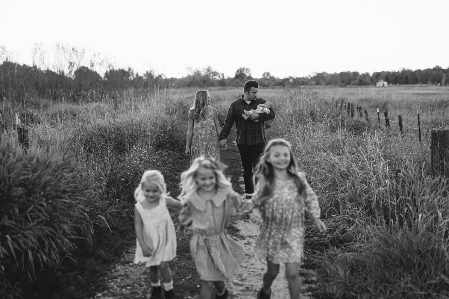 Three children run joyfully through a tall grass field while adults follow behind them.