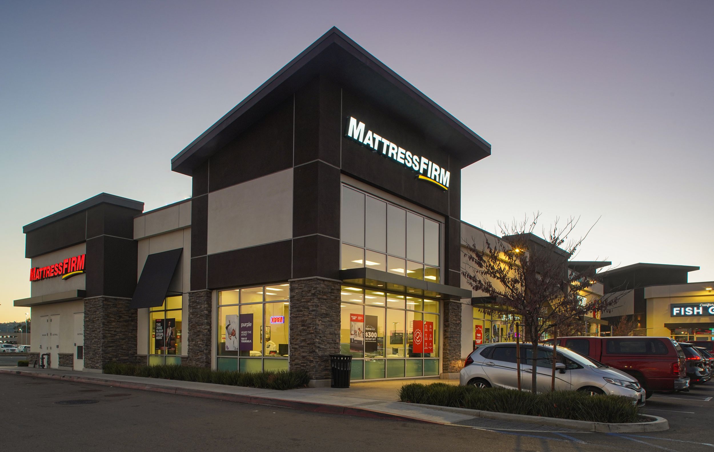 Brightly lit Mattress Firm retail store exterior with large glass windows and parking in a commercial shopping plaza.