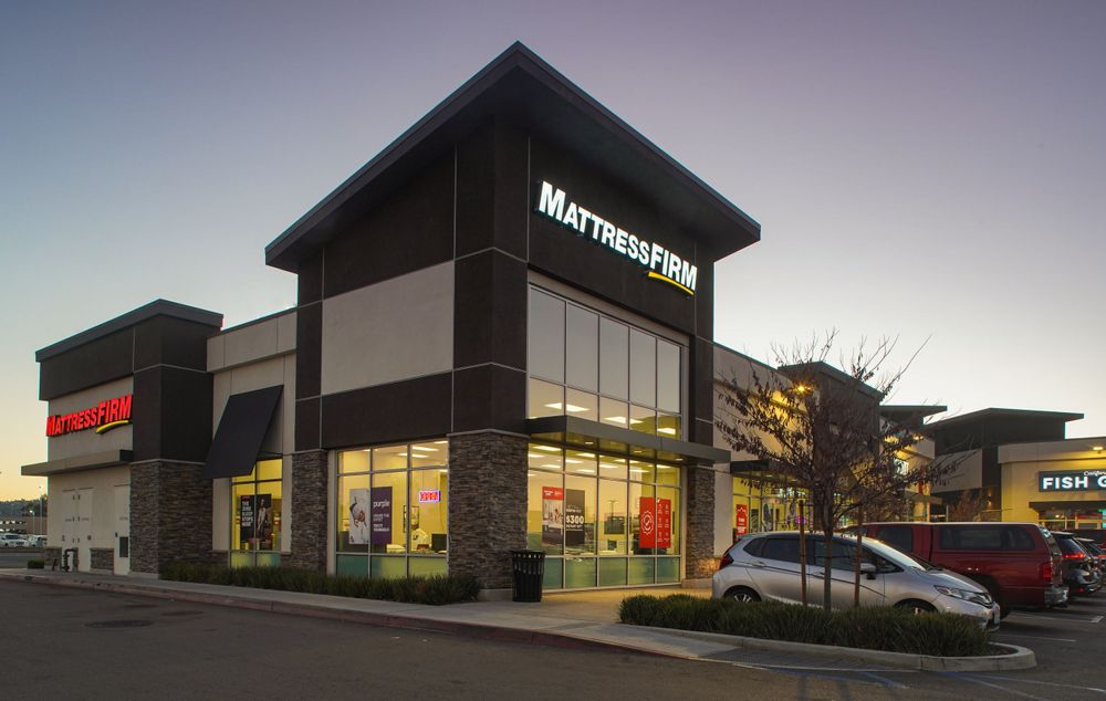 Brightly lit Mattress Firm retail store exterior with large glass windows and parking in a commercial shopping plaza.