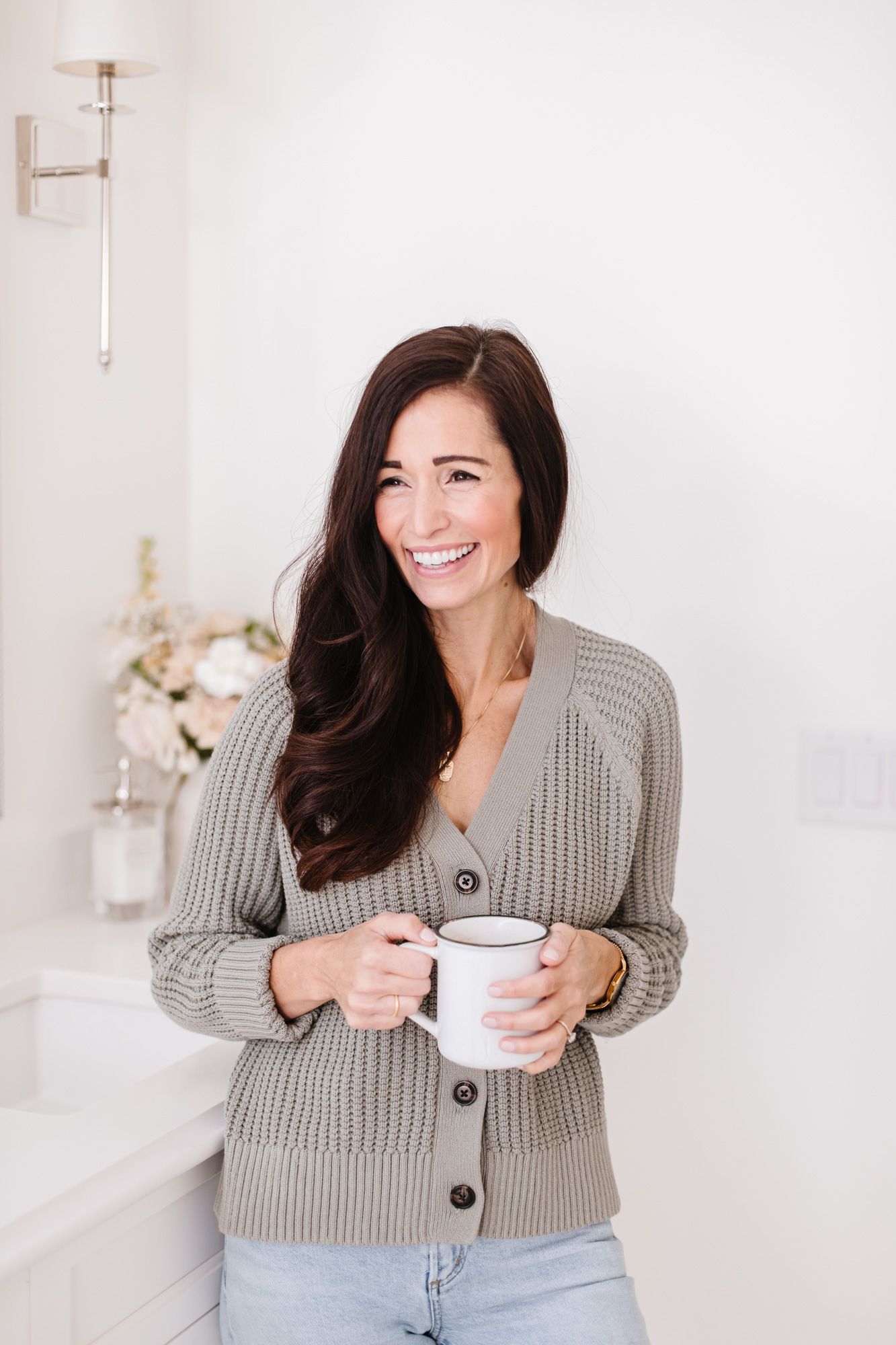 Woman in gray knit cardigan holding white coffee mug against light background.