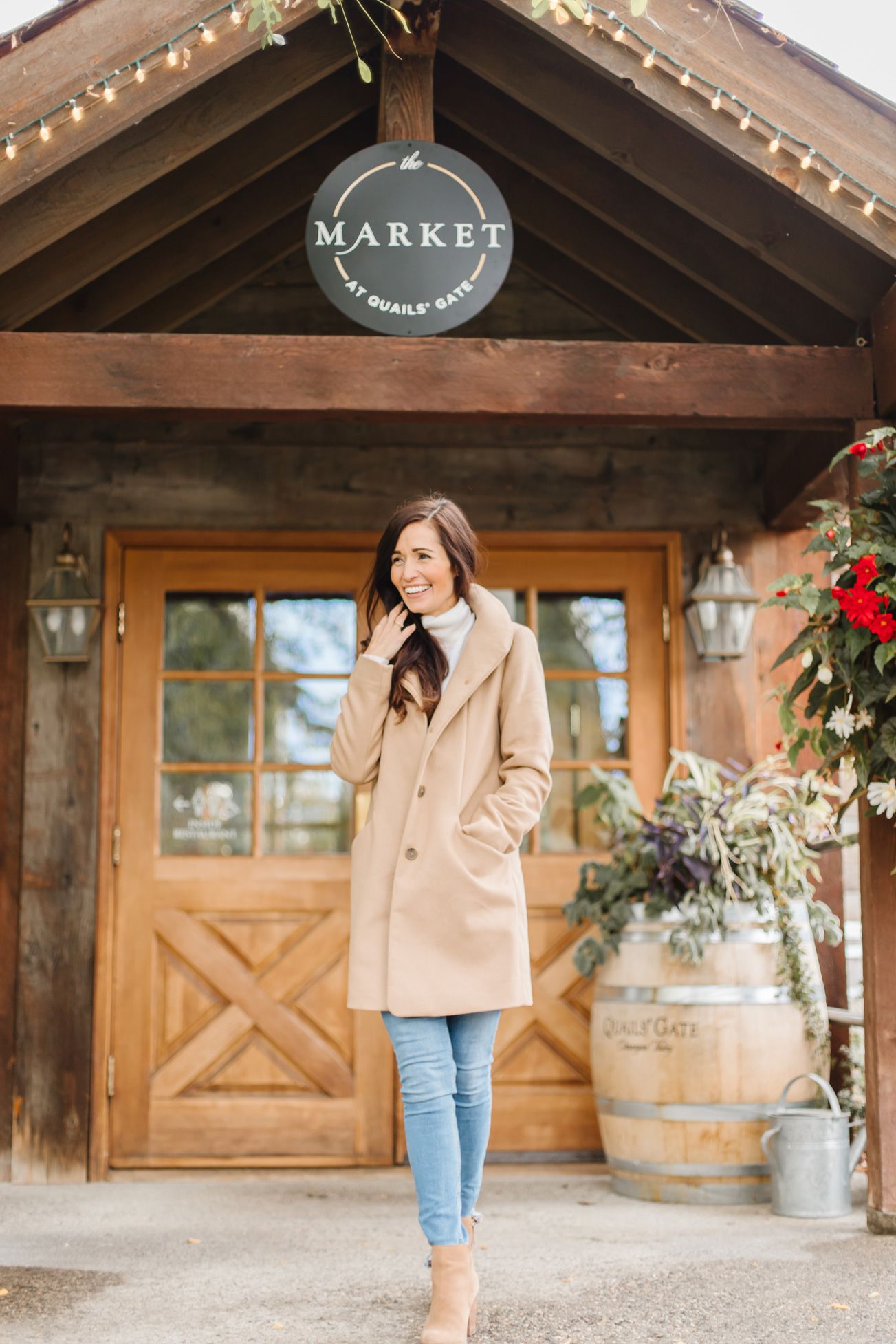 Stylish camel coat and jeans outfit in front of rustic market entrance with string lights.