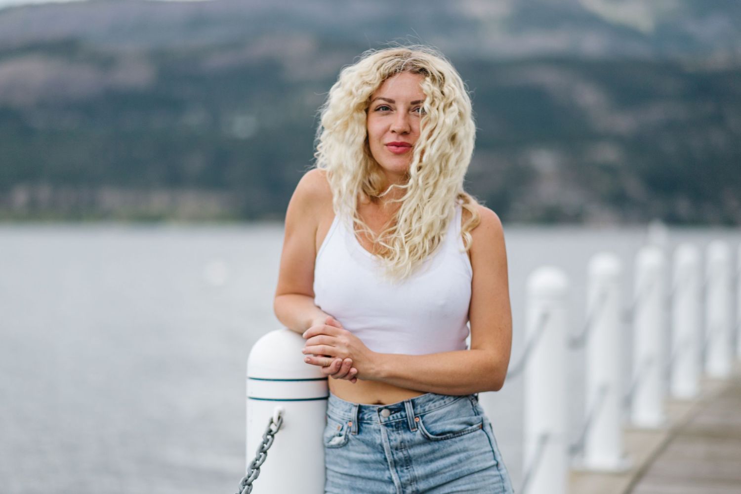 Person in casual attire standing near waterfront with mountains in background.