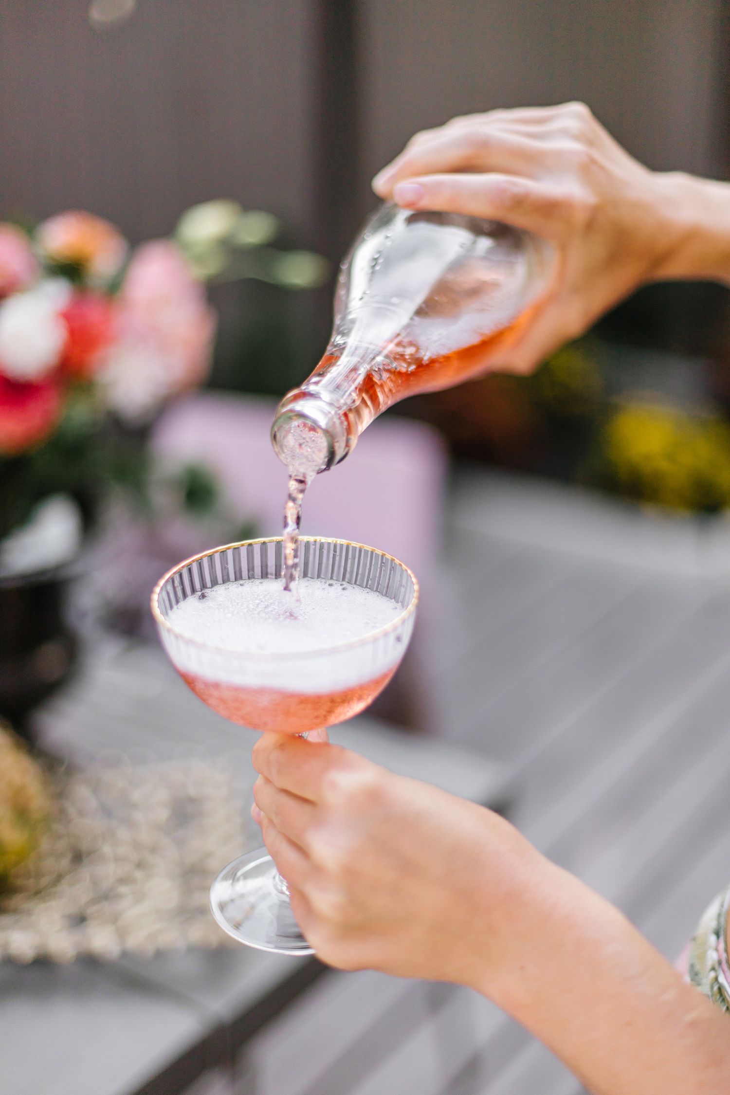 Pink cocktail being poured into a coupe glass with flowers in the background.