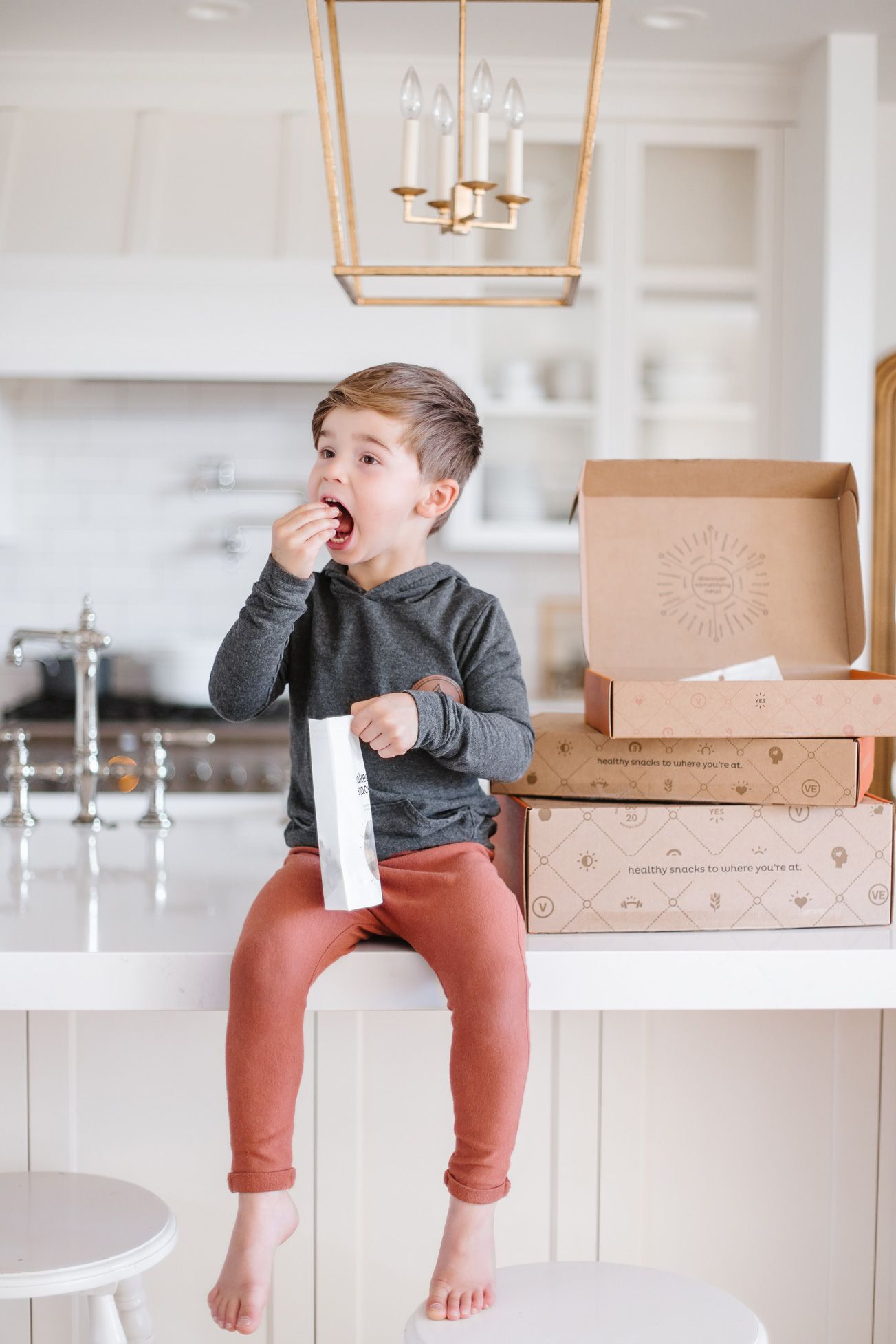 Child in gray sweater and rust-colored pants sits playfully on white kitchen counter with cardboard boxes.