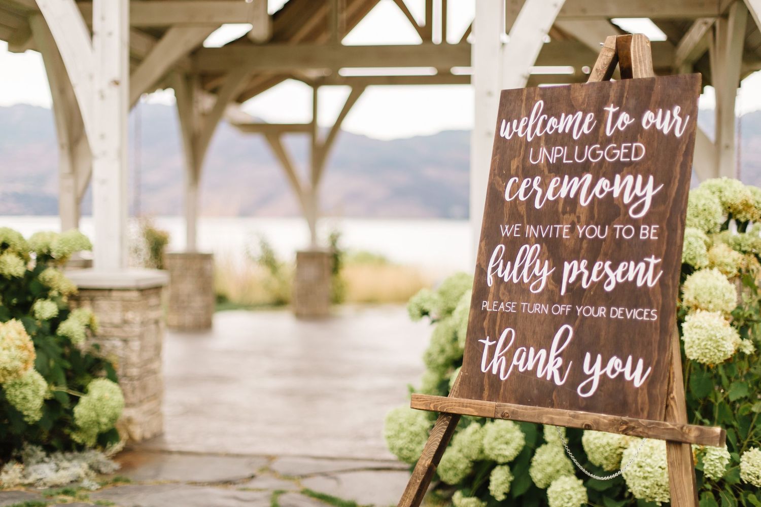 Wooden unplugged ceremony welcome sign displayed on an easel at an outdoor venue.