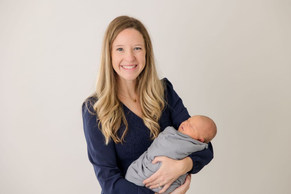 A woman in navy blue holds a swaddled newborn baby against a light colored background.