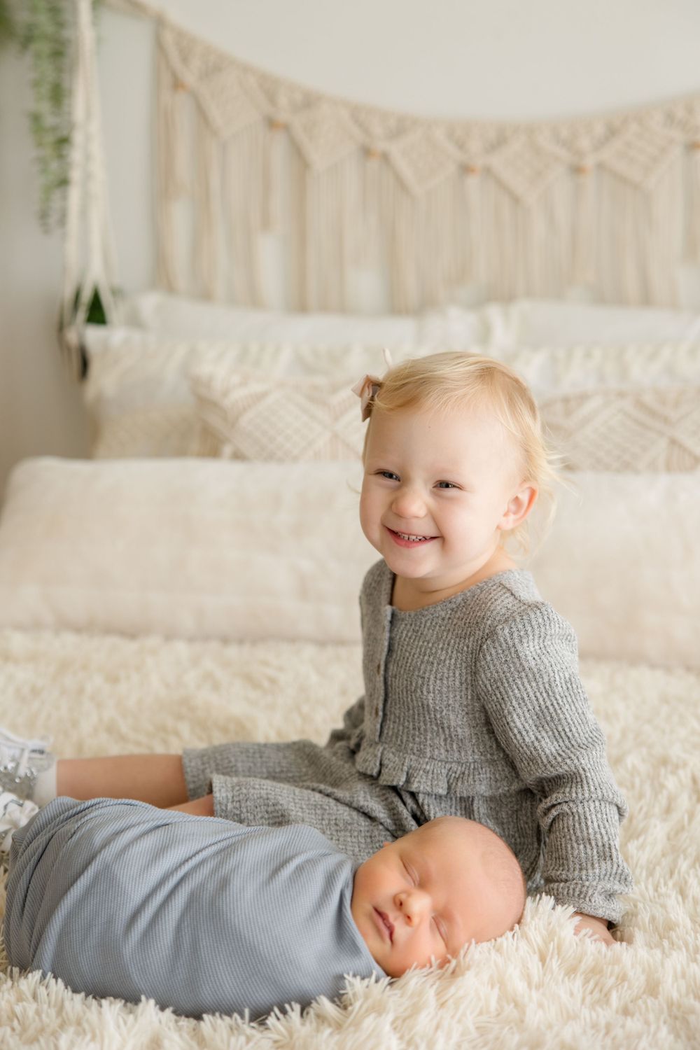 Two siblings sharing a sweet moment on a white bed with macrame wall decor.