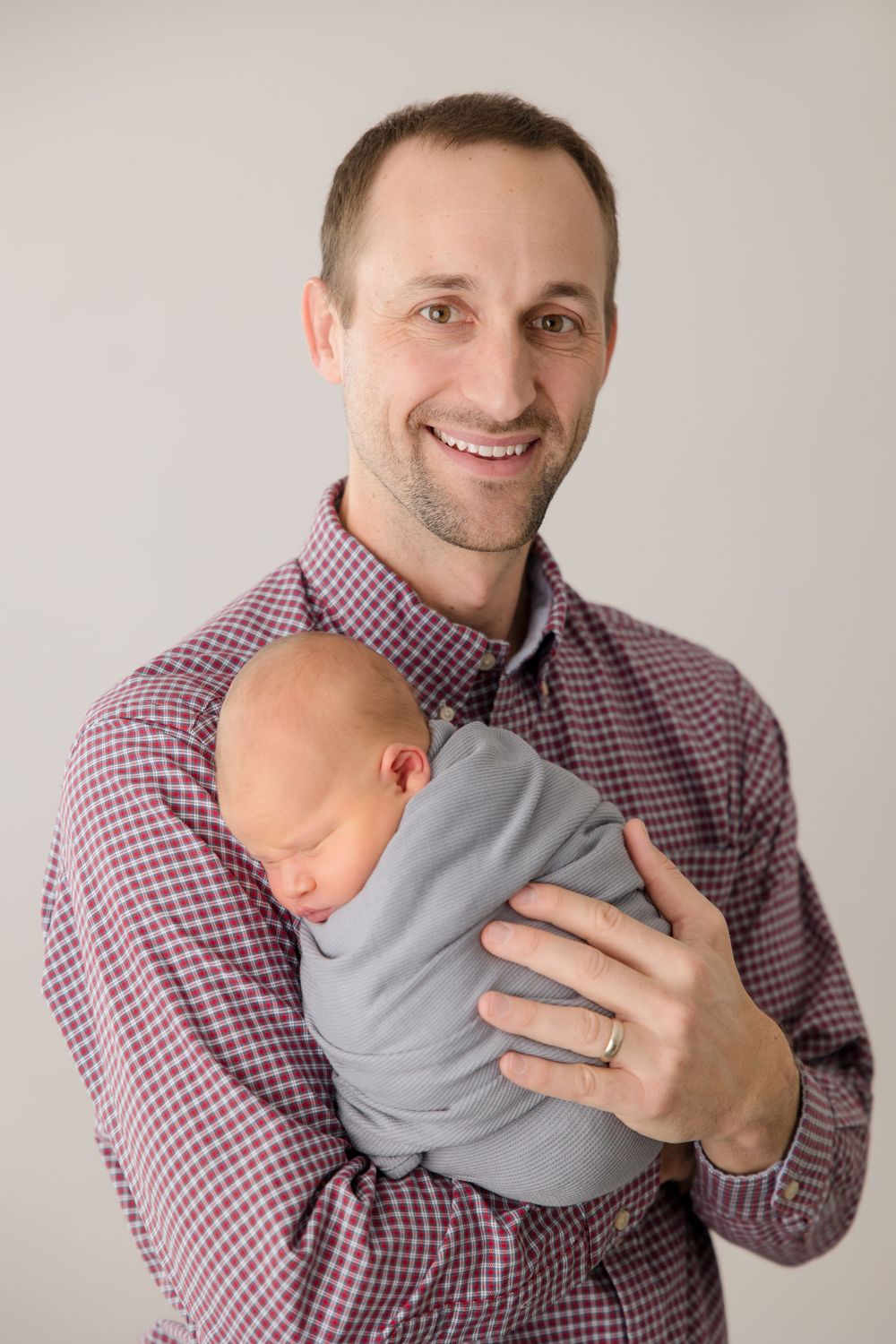 A man in a checkered shirt cradles a sleeping newborn wrapped in gray.