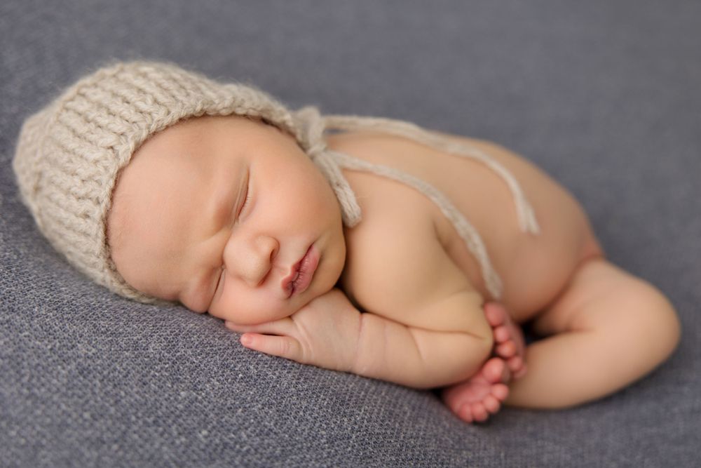 Sleeping newborn baby wearing knit hat curled up on gray blanket in peaceful studio portrait.