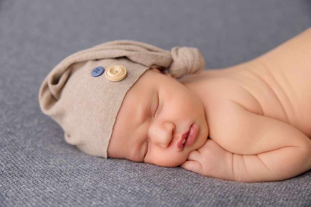 Peaceful newborn sleeps while wearing a tan knit cap with buttons on a gray background.