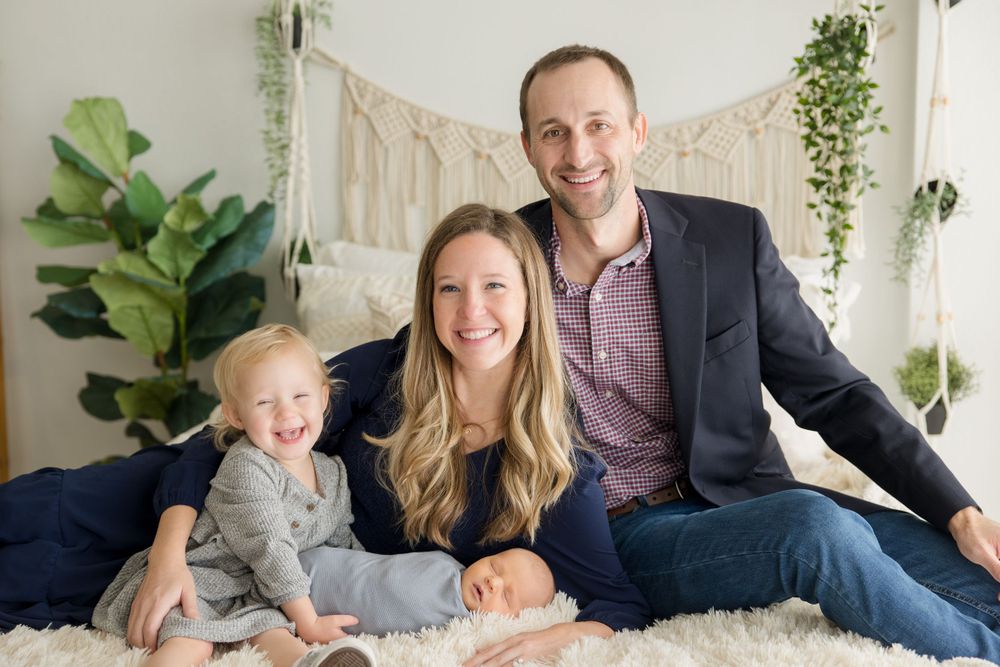 Family portrait on white bedding with macrame wall hanging and indoor plants.