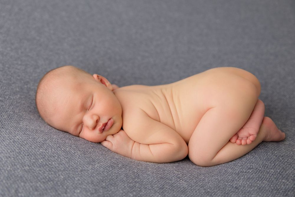 A sleeping newborn baby lies curled up on a gray backdrop in a natural pose.