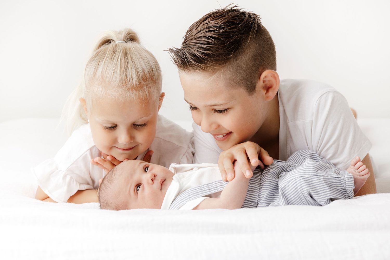 Siblings sharing a sweet bonding moment with a newborn baby on white bedding.