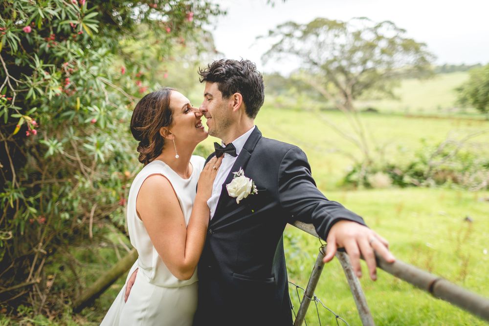Couple in formal wedding attire embrace near rustic fence in countryside setting.
