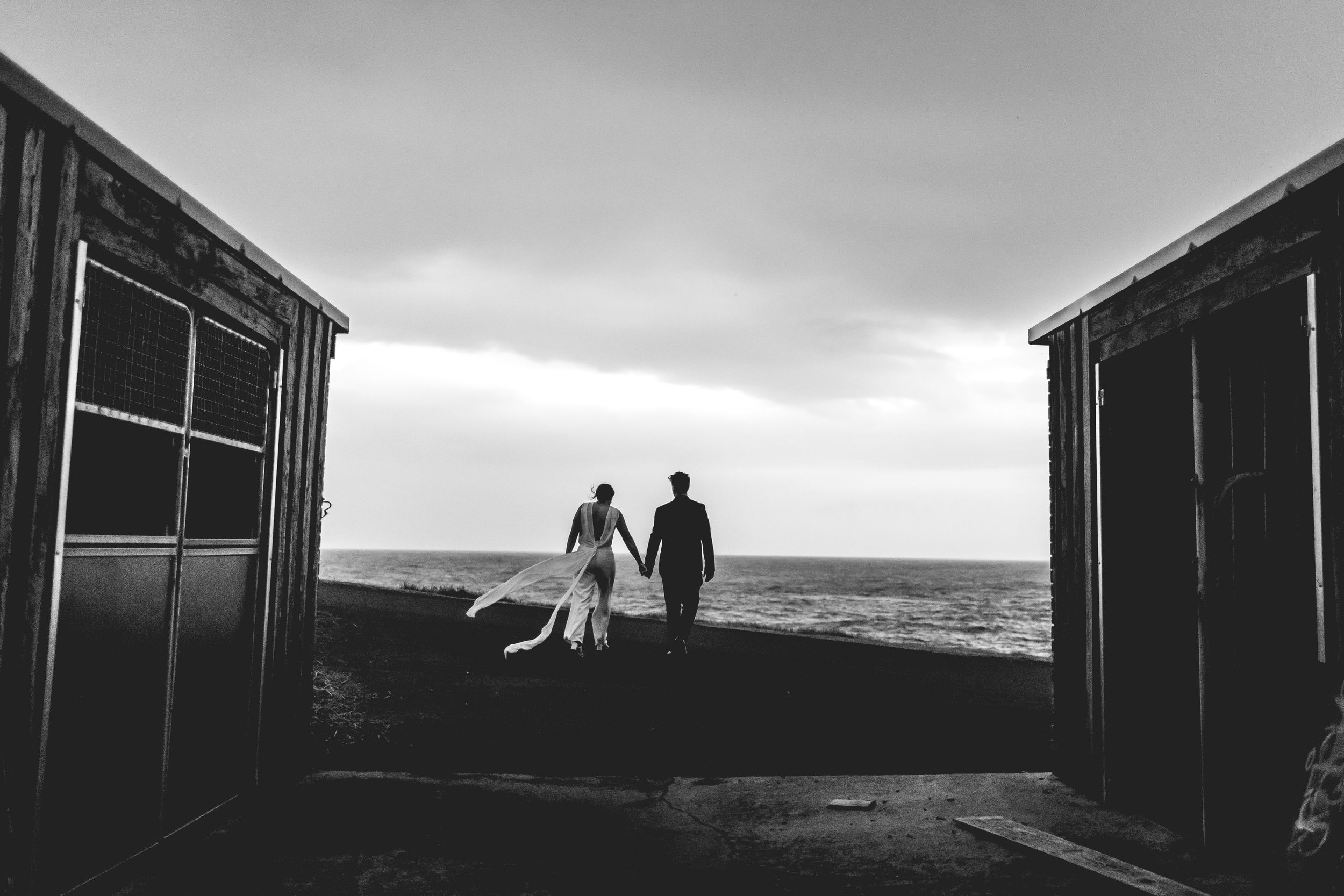 Black and white silhouette of couple walking between buildings toward ocean horizon.