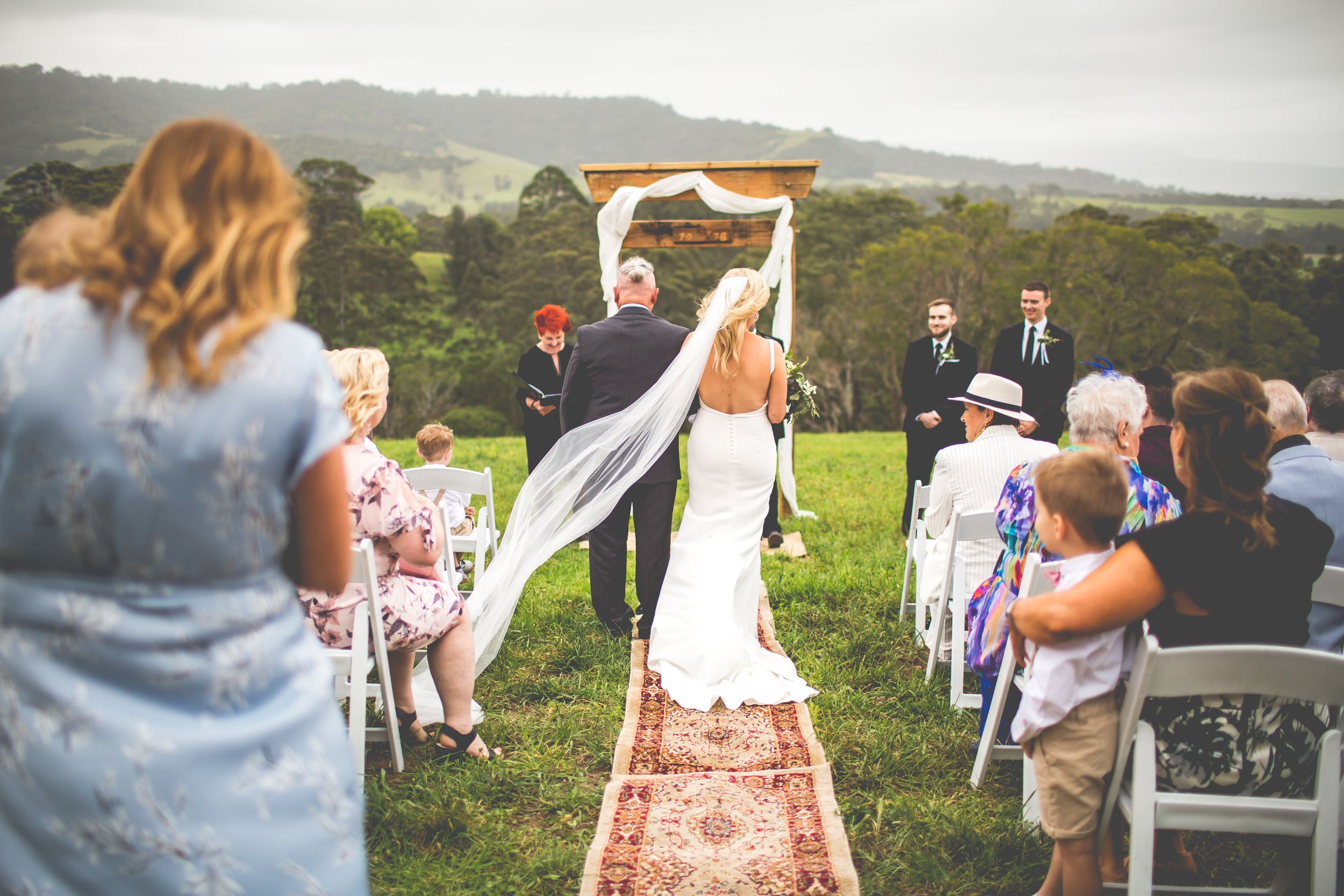 Outdoor wedding ceremony setup with decorated aisle runner and chairs on grassy hillside.