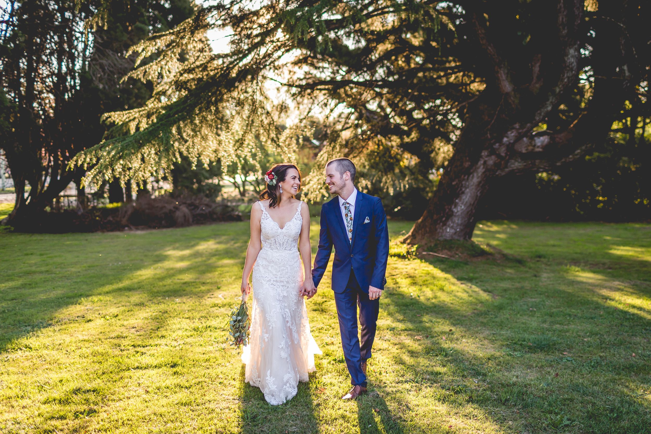 A couple walks hand in hand through a sunlit garden with golden rays streaming through tree branches.