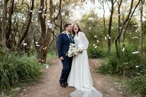 A couple shares a romantic moment on a woodland path while holding a white floral bouquet.