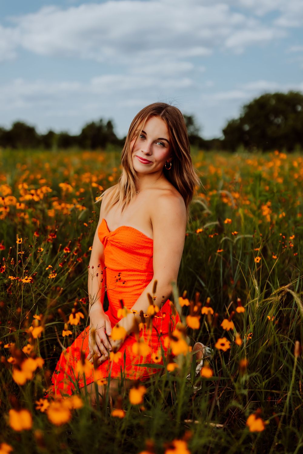 Woman in orange dress sits among vibrant orange wildflowers under blue summer sky.