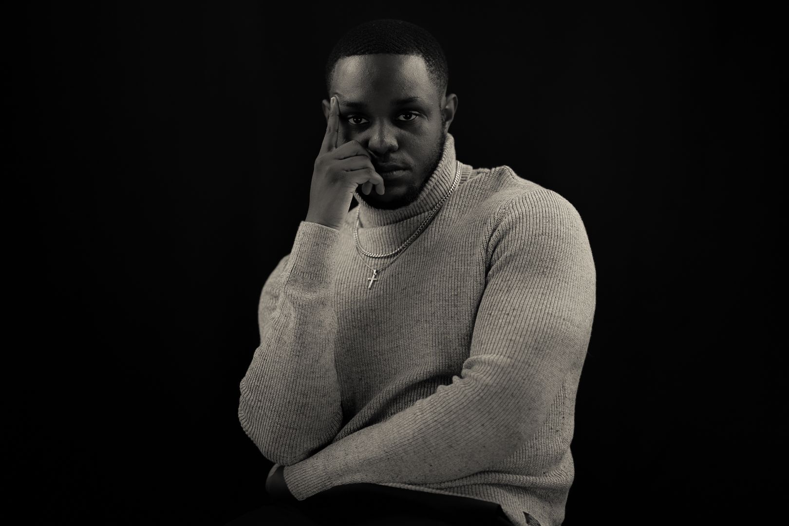 A person in light-colored sweater poses thoughtfully with hand on face against dark background.