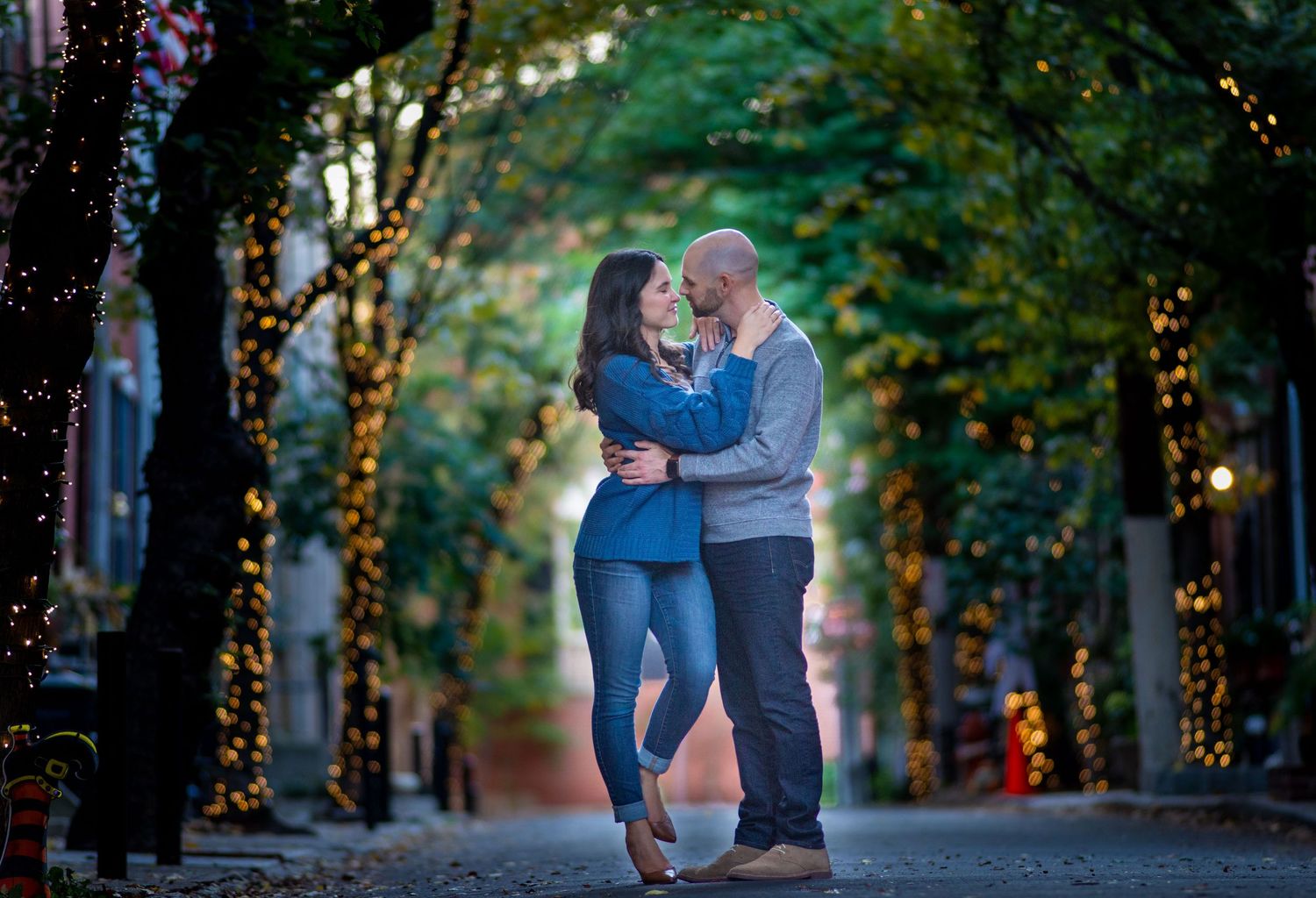A romantic couple embraces on a pathway lined with twinkling lights and trees in a magical garden setting.