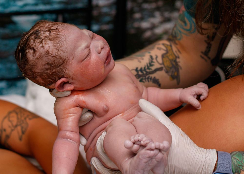 Close-up photo of newborn baby being held by caregiver with visible tattoos.