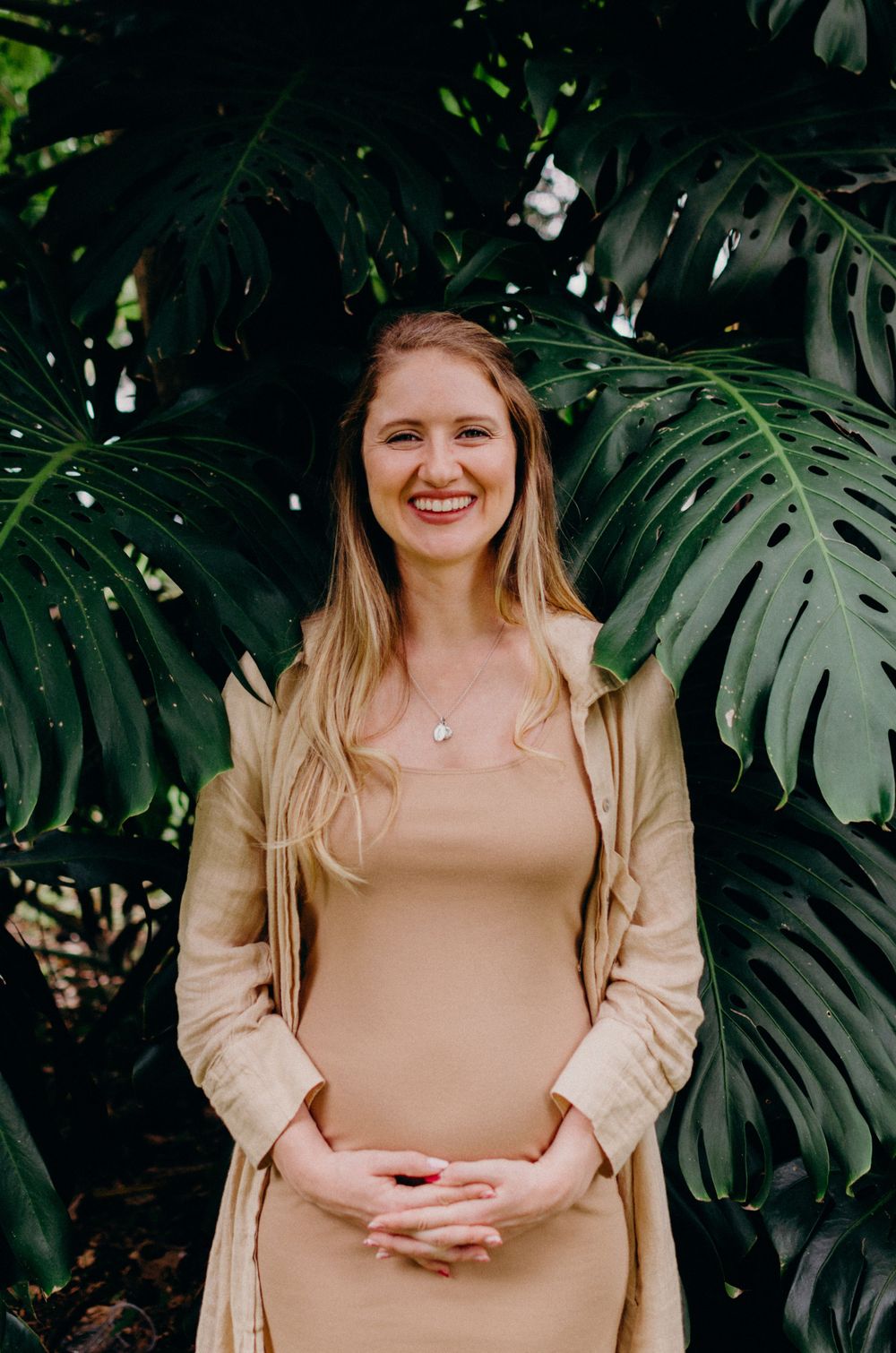 Woman in beige dress smiling while posing against large monstera leaves.