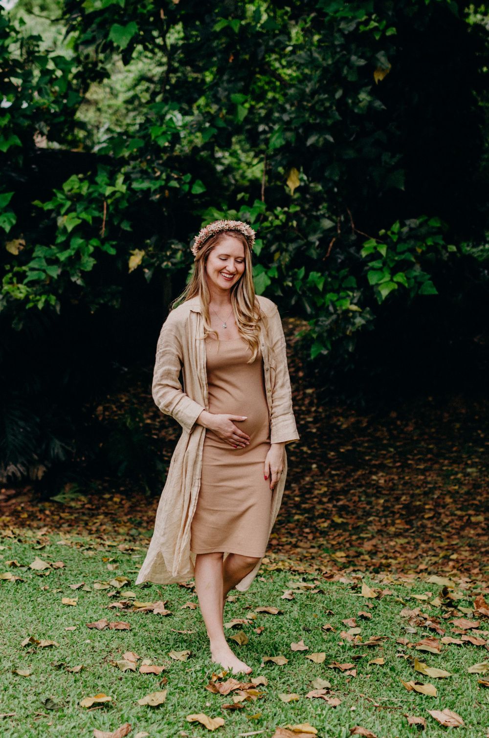 Barefoot maternity portrait on grass with autumn leaves and a flowing beige ensemble.