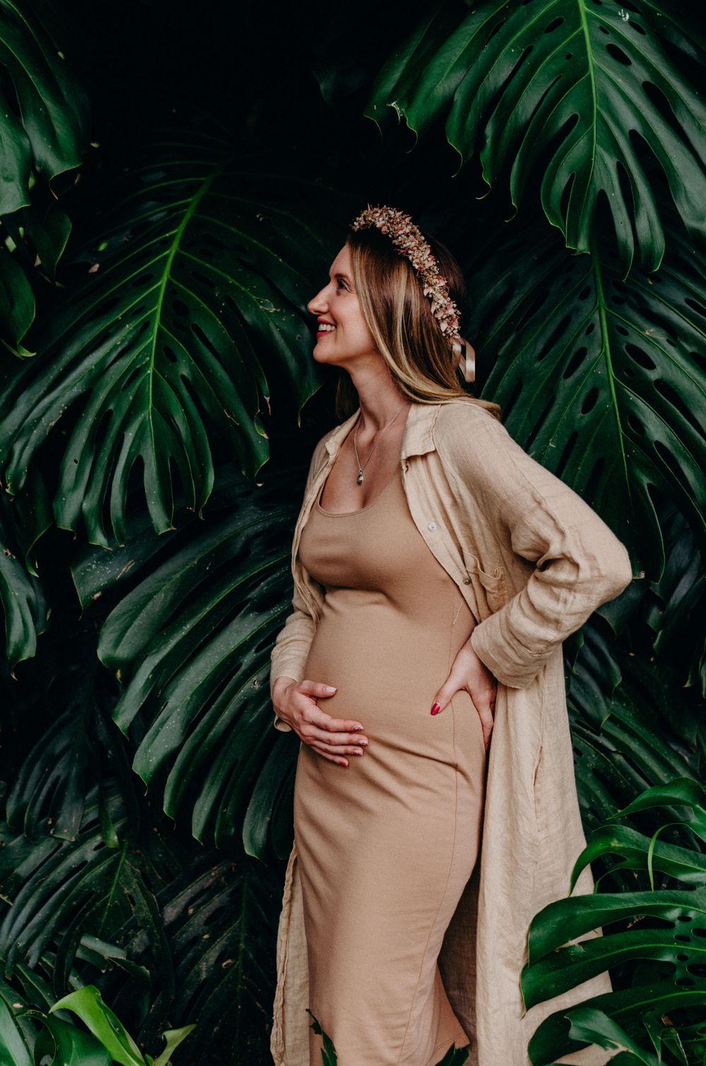 Elegant maternity photo against tropical monstera leaves wearing beige fitted dress.