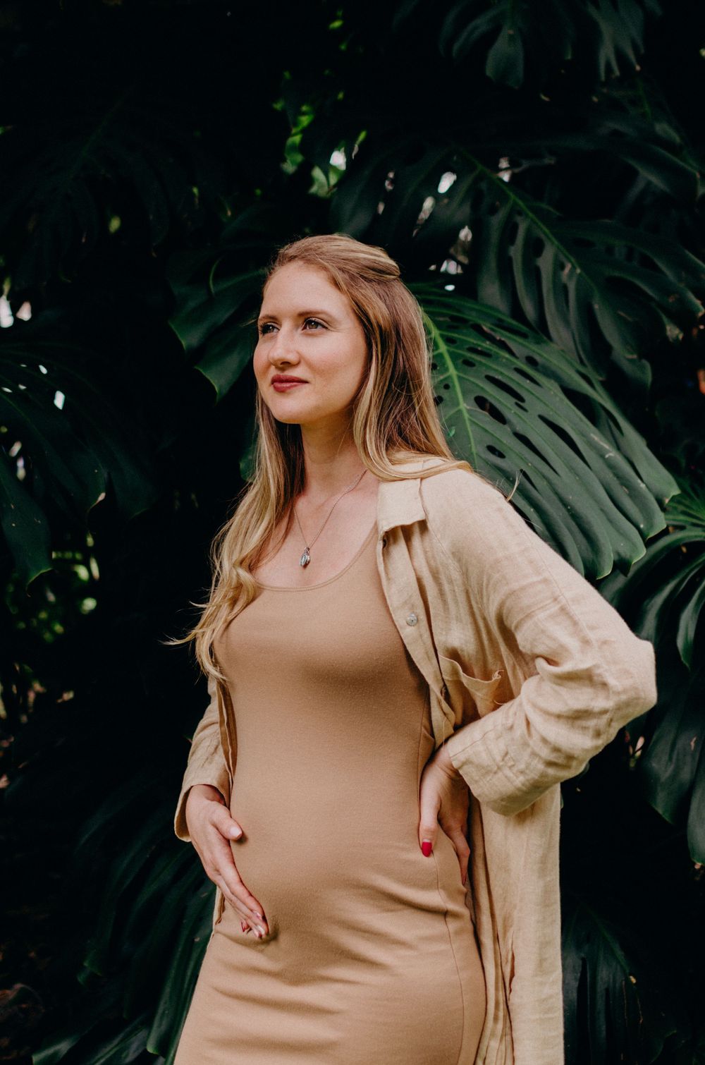 Expectant mother in beige dress and cardigan poses against tropical foliage.