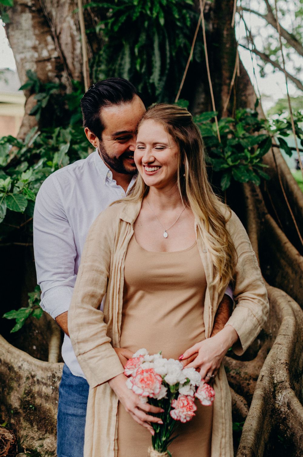 Couple embracing near a large tree while holding pink flowers.