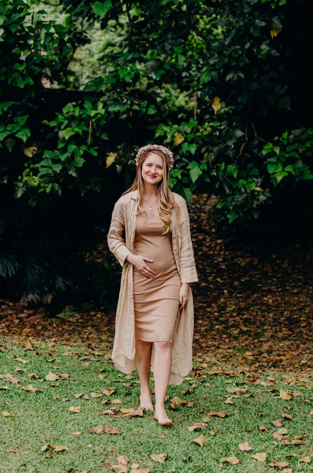 A maternity photoshoot in a garden setting showing a beige dress and long cardigan against lush greenery.