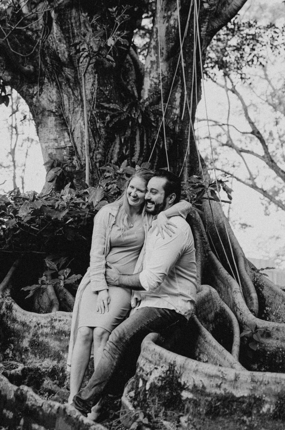 Black and white photo of a couple sitting on tree roots in a natural setting.