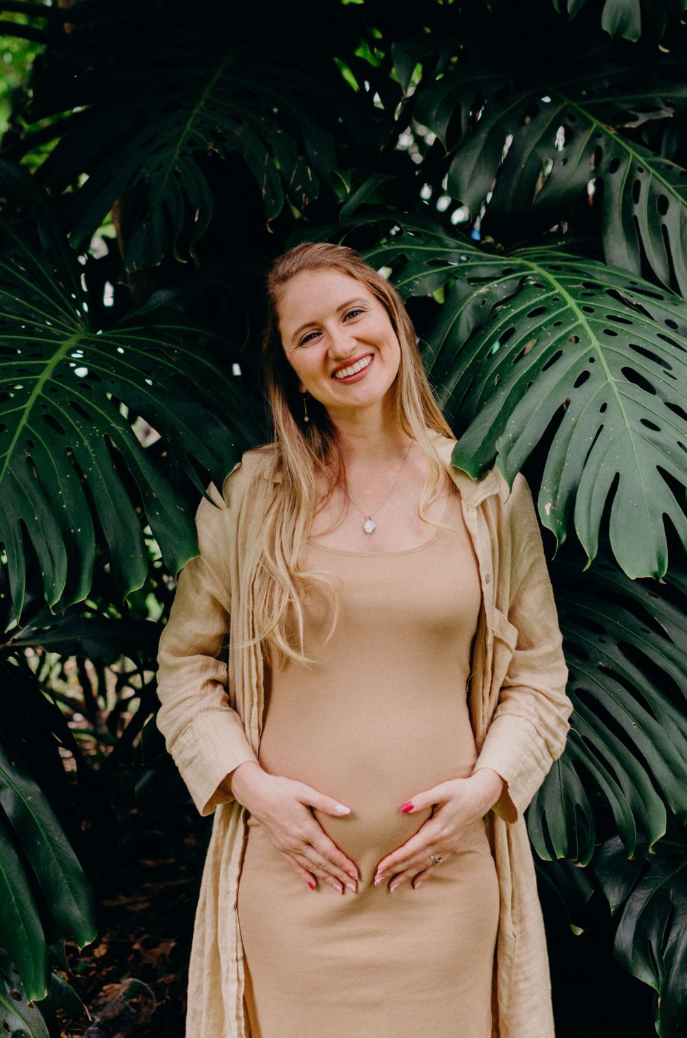 Smiling woman in neutral tones standing against tropical plant backdrop.