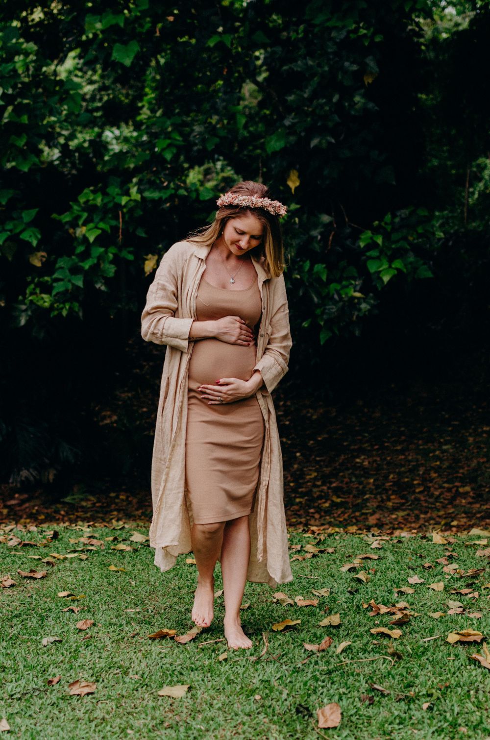 Expectant mother wearing floral crown and beige outfit stands barefoot on autumn grass.
