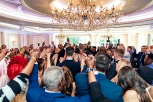 Crowd of wedding guests dancing under crystal chandelier in elegant ballroom reception.